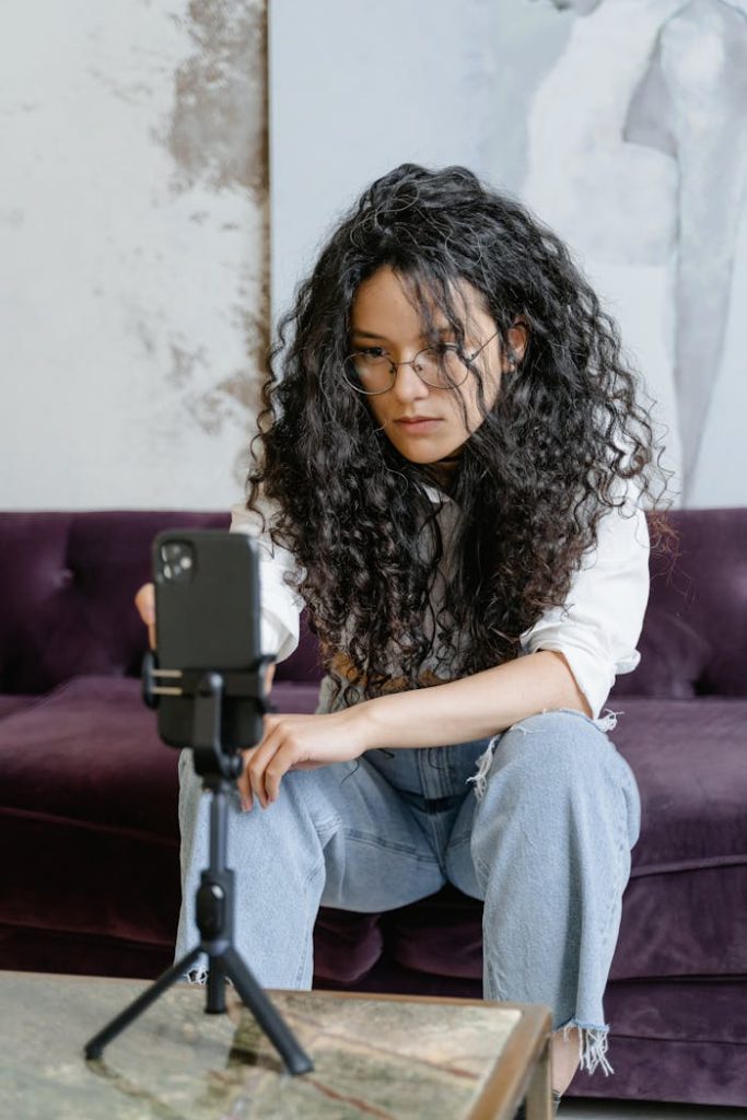 Woman with curly hair vlogging indoors, using smartphone on tripod, seated on couch.